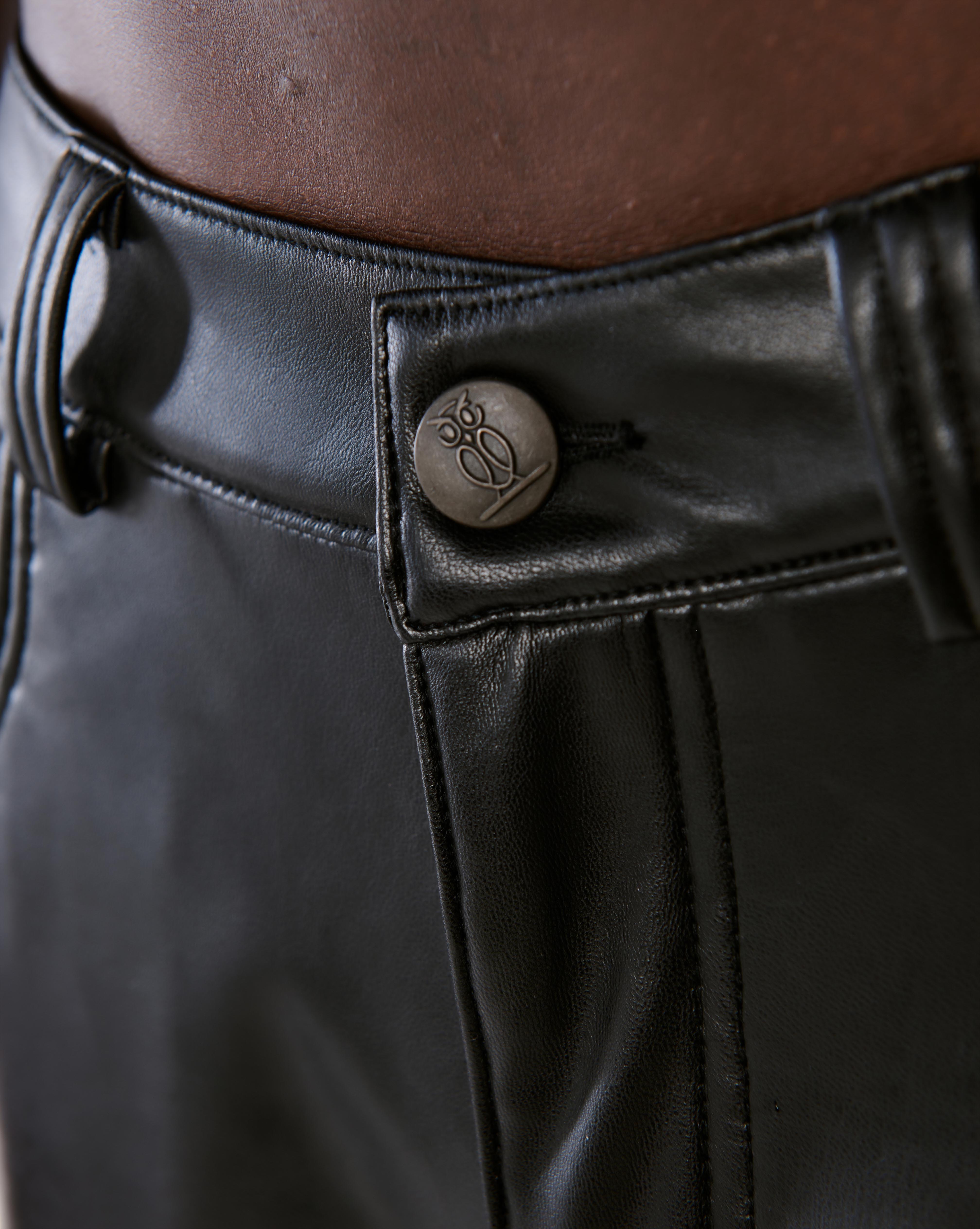 Detailaufnahme des gravierten Metallknopfes mit MOE CHARLIE Owl-Logo am Hosenbund der schwarzen Lederhose