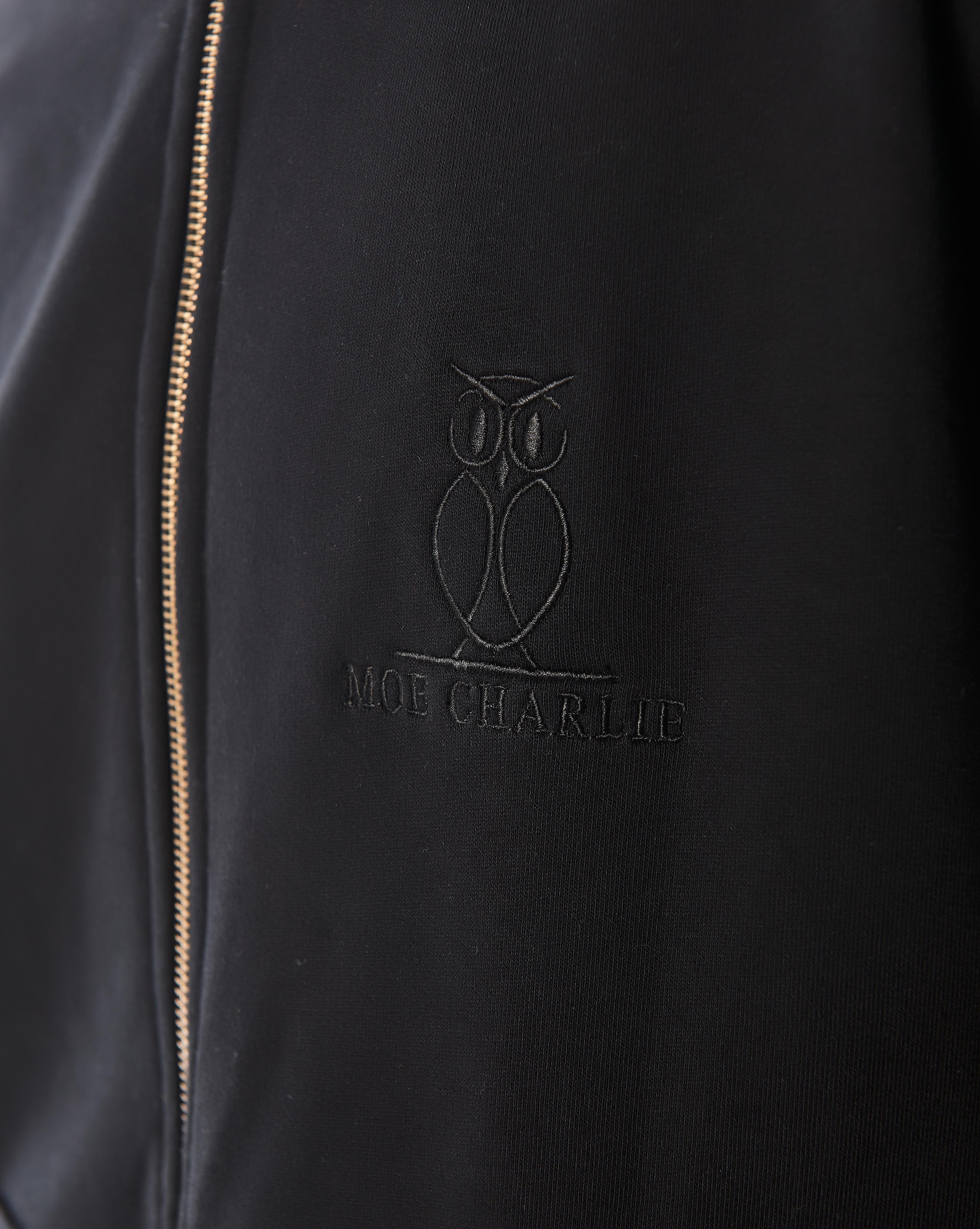 Makroaufnahme des schwarz-gestickten MOE CHARLIE Owl-Logos und Schriftzugs ton-in-ton auf der schwarzen Zip-Jacke