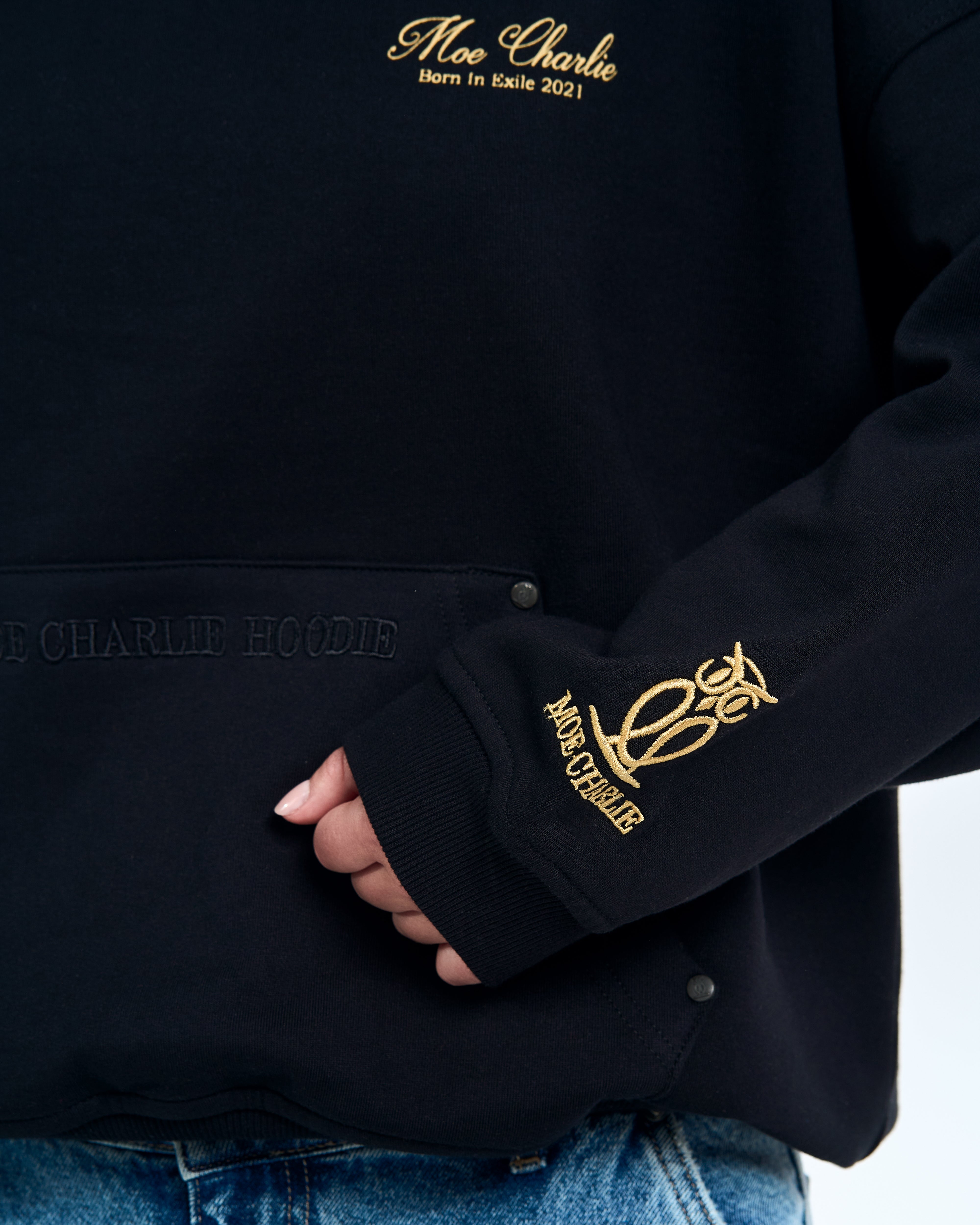Detailaufnahme MOE CHARLIE HOODIE Schriftstickerei auf schwarzem Stoff, Taschenkante und Druckknopf