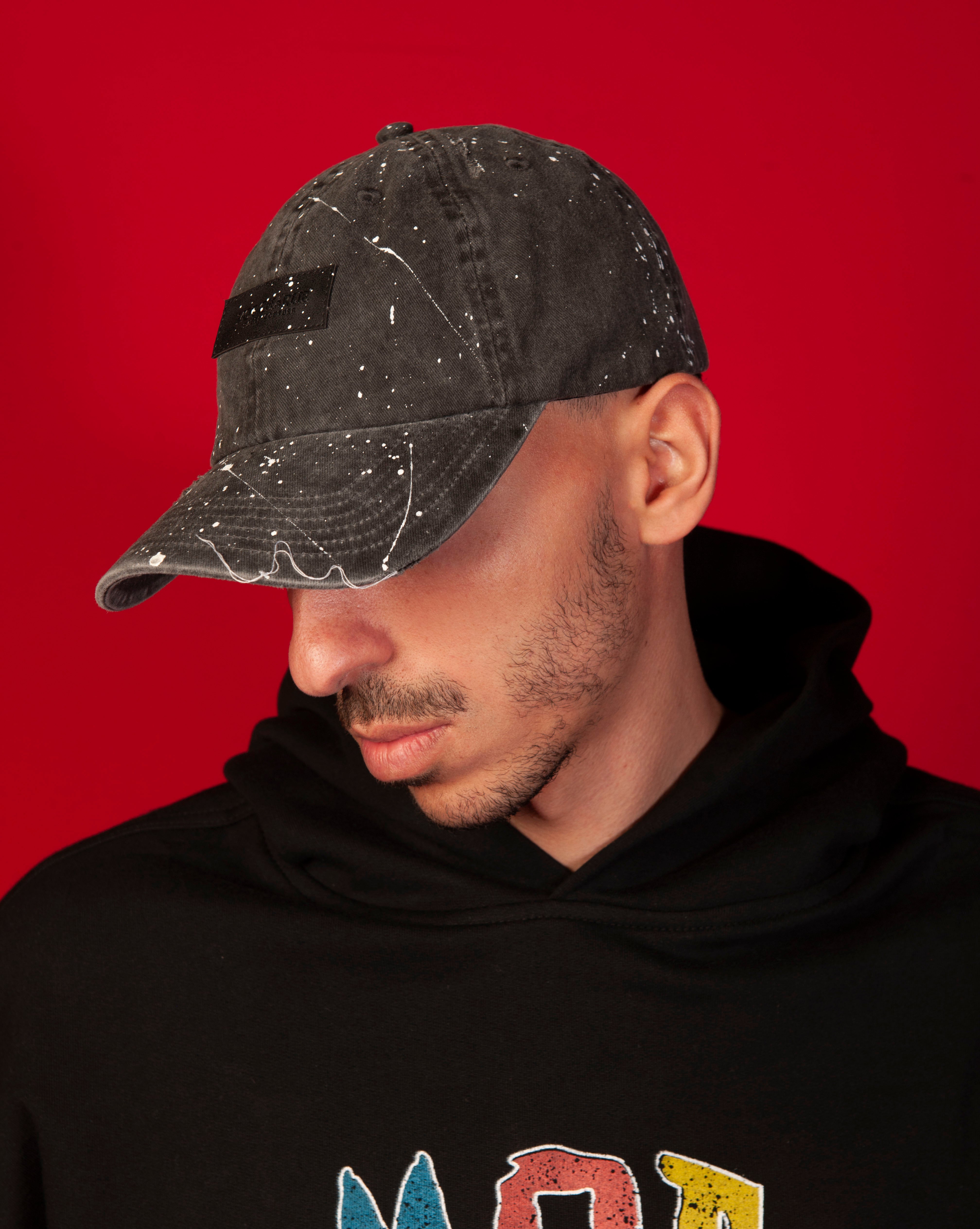 Destroyed MOE CHARLIE Cap, Seitenportrait maennlich, schwarzes Paint-Splatter-Cap mit Label-Patch, Blick nach unten