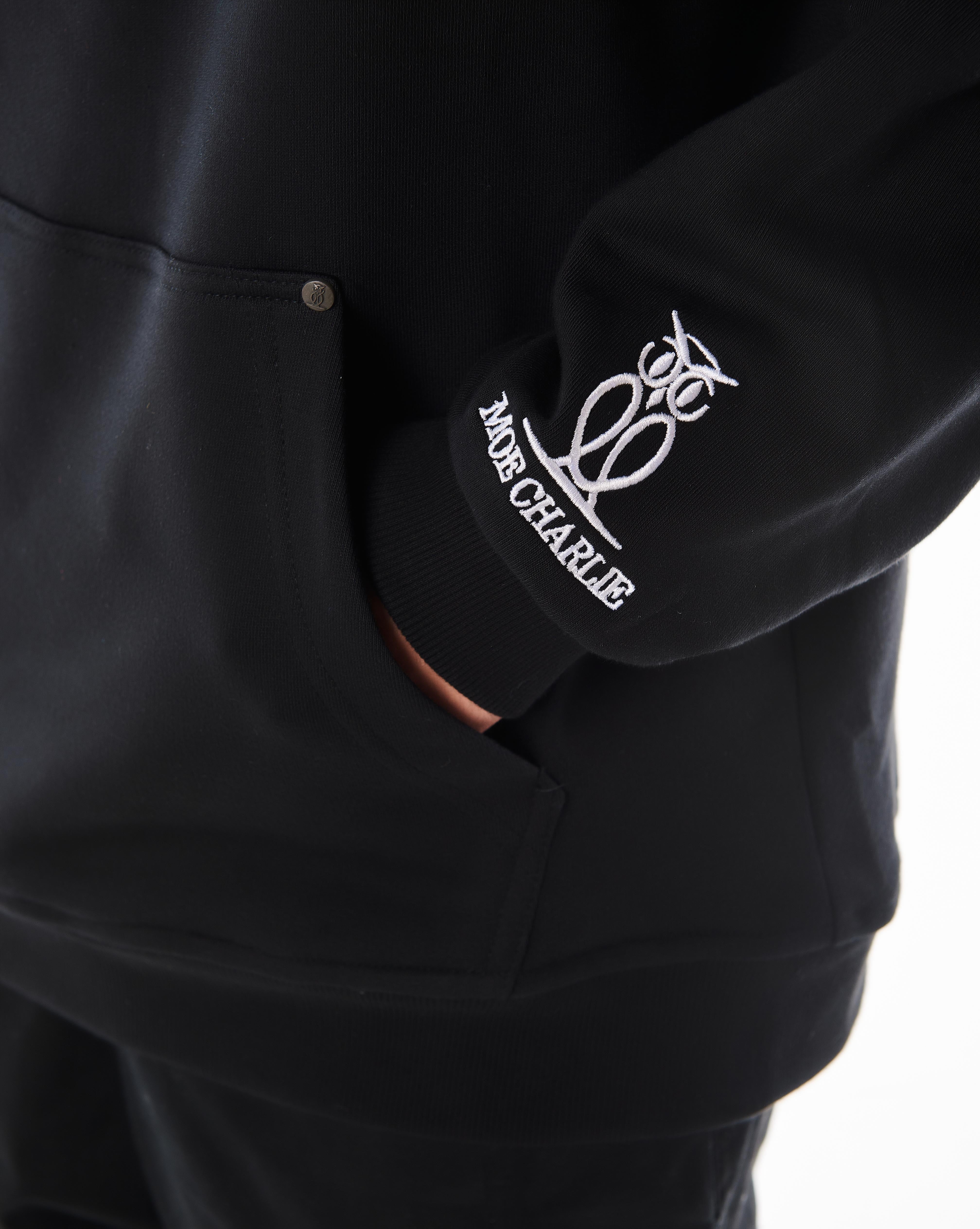 Detailaufnahme Ärmelstickerei MOE CHARLIE Predator Hoodie, Eulen-Logo am Bündchen, Hand in Känguru-Tasche