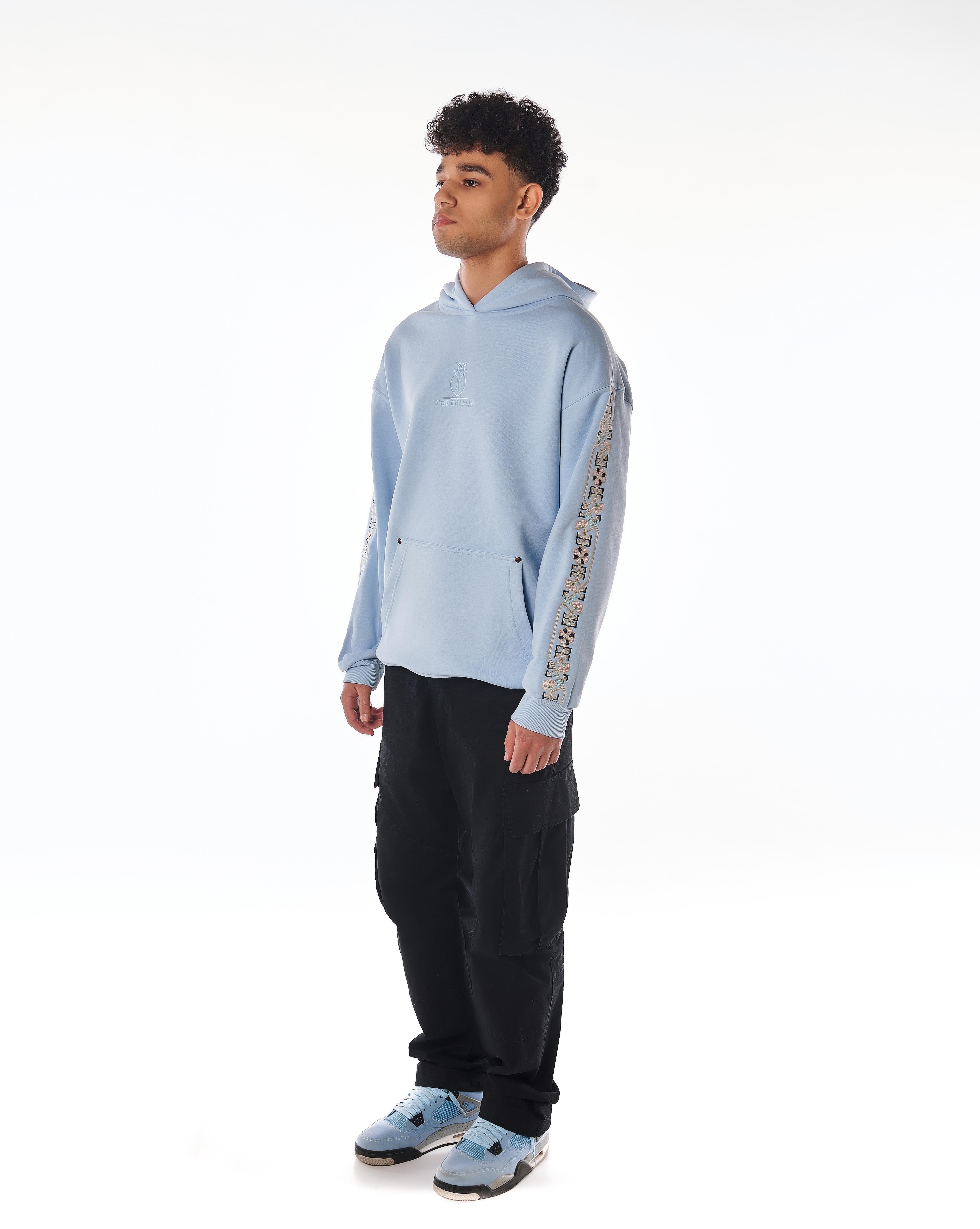The Culture Hoodie Riviera Blue von MOE CHARLIE, Ganzkörperpose von links, florale Ärmelstickerei, Cargohose und Sneaker