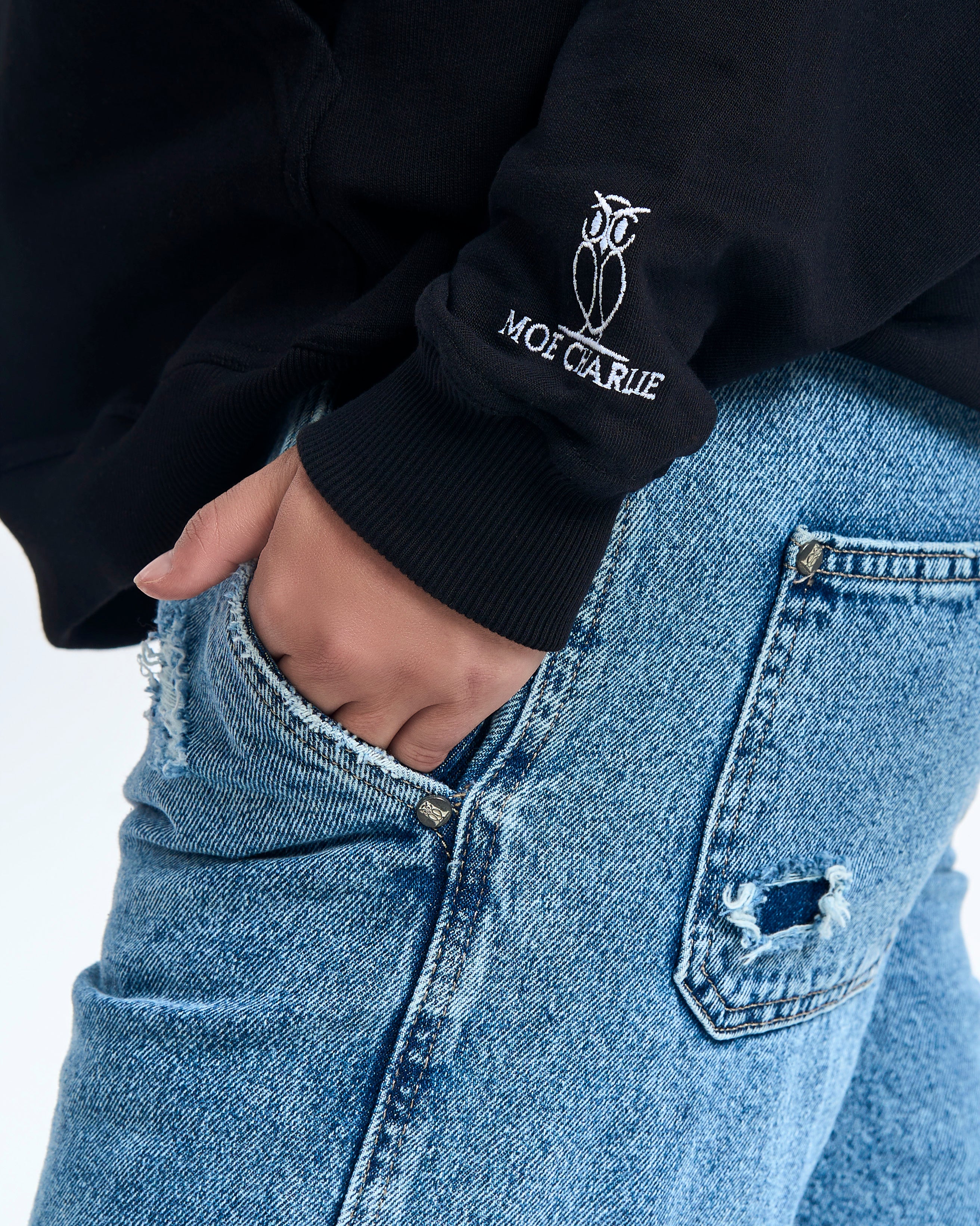 Aermel-Nahaufnahme MOE CHARLIE Hoodie Schwarz - Owl-Crest-Stickerei am Buendchen, Hand in Jeans-Tasche