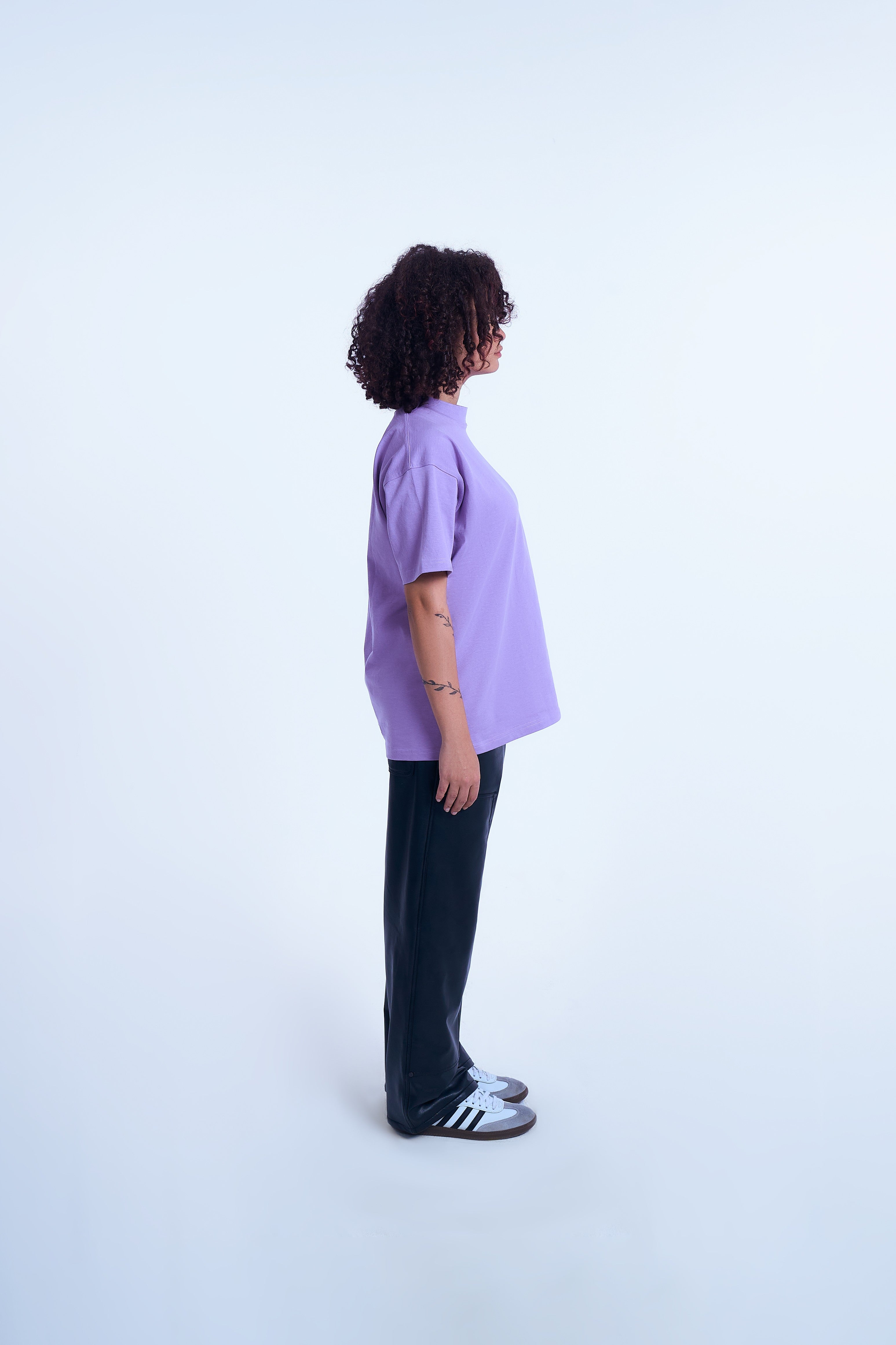 Seitenansicht weiblicher Model MOE CHARLIE Lavendel Basic Tee – schwarze Lederhose, Adidas Samba