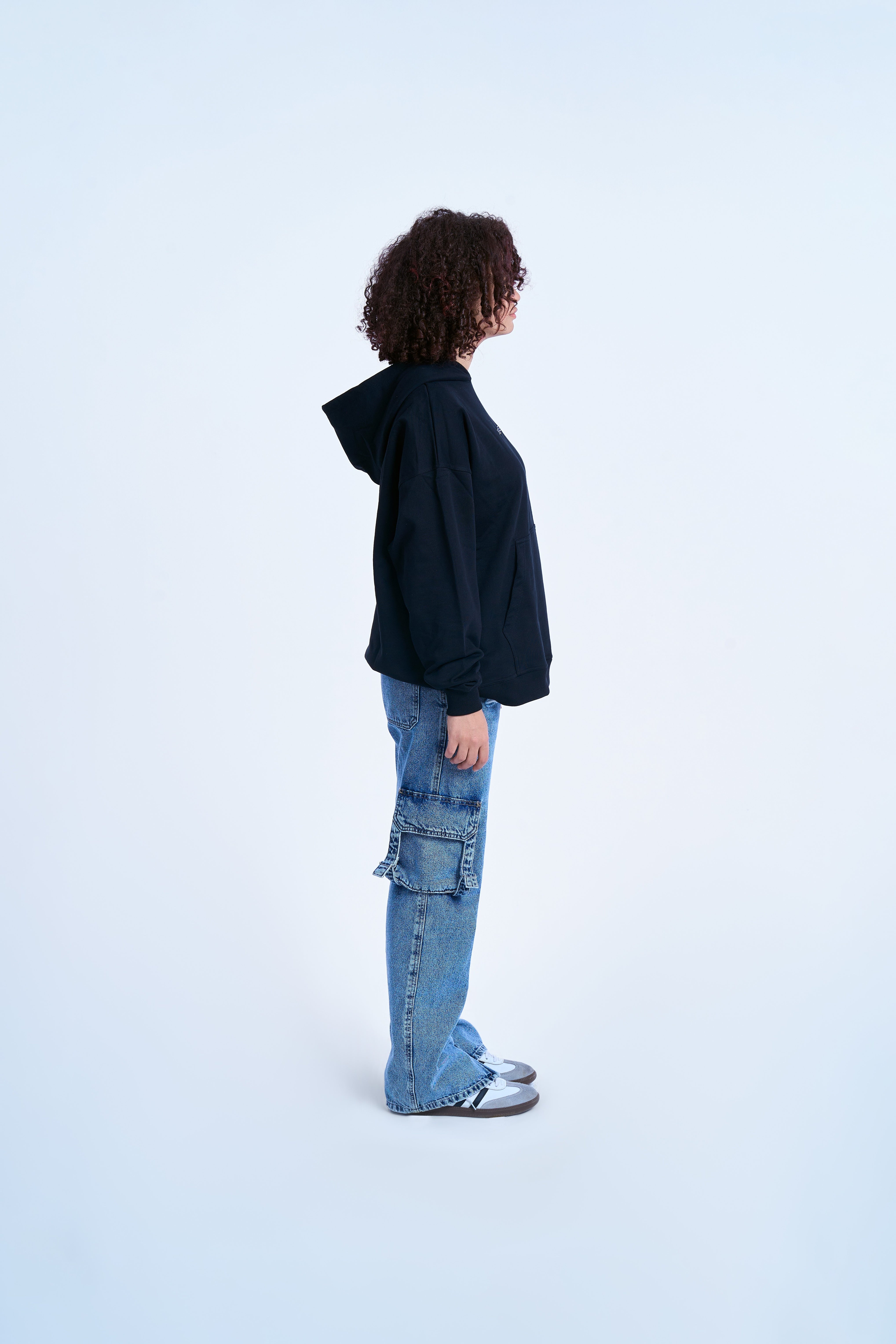 Schwarzer MOE CHARLIE Hoodie, weibliche Traegerin, Seitenansicht mit Kapuze, Cargo-Jeans und Sneakers