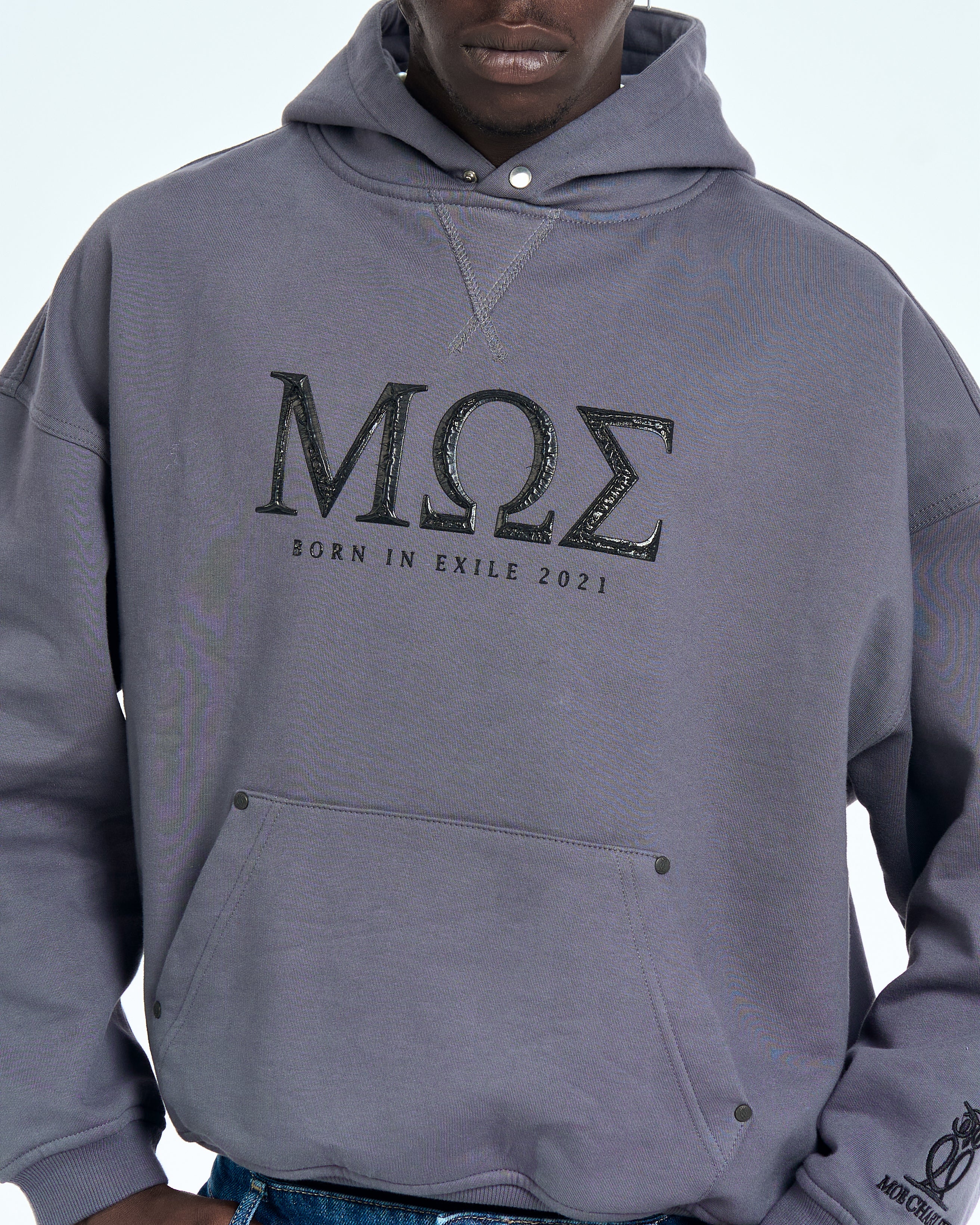 Nahaufnahme MOE CHARLIE Born in Exile Hoodie Grau, Brustbereich mit glossy MOE-Print und Druckknopfen
