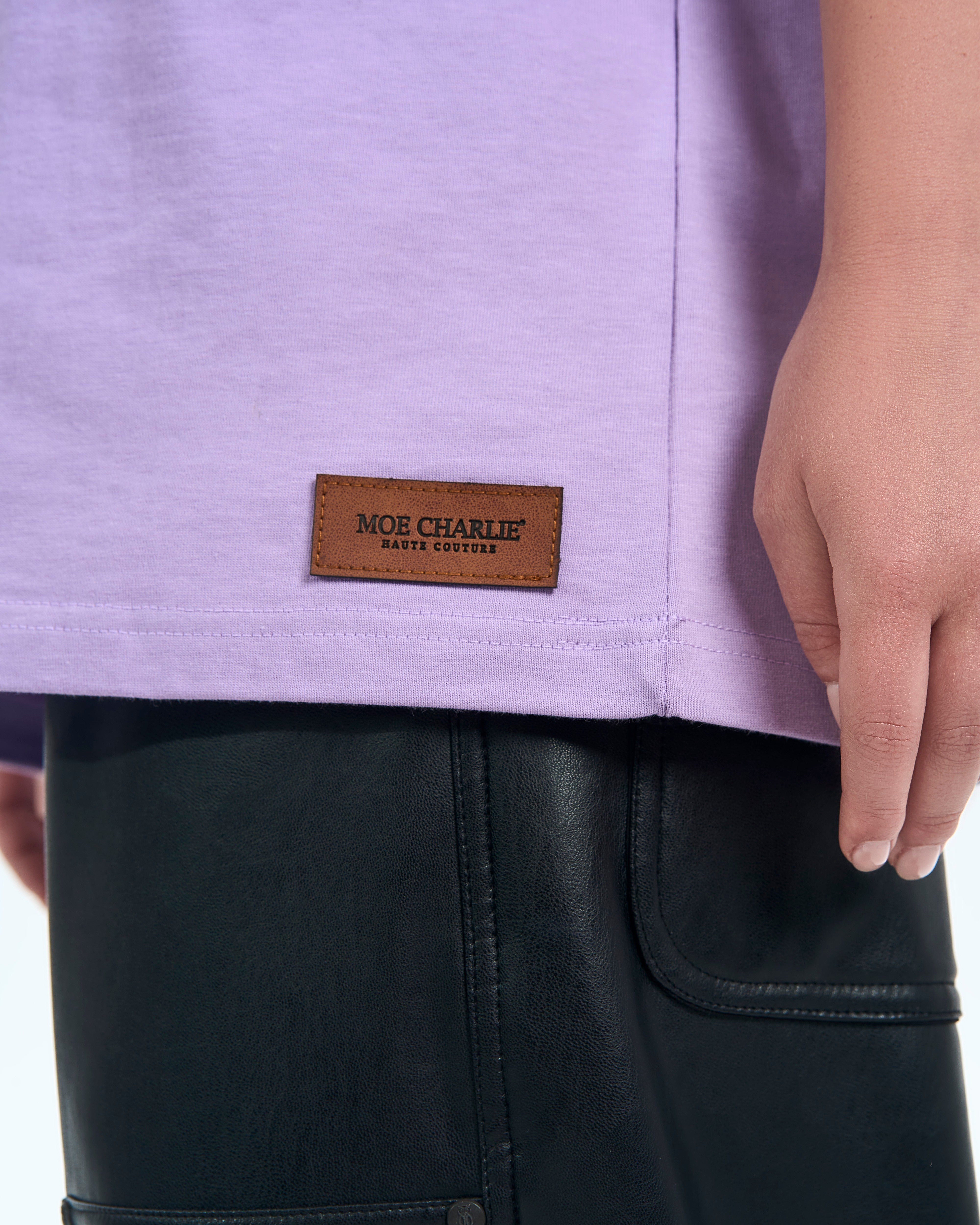 Nahaufnahme Lederpatch Detail am Saum – MOE CHARLIE Haute Couture Etikett auf lavendelfarbenem T-Shirt