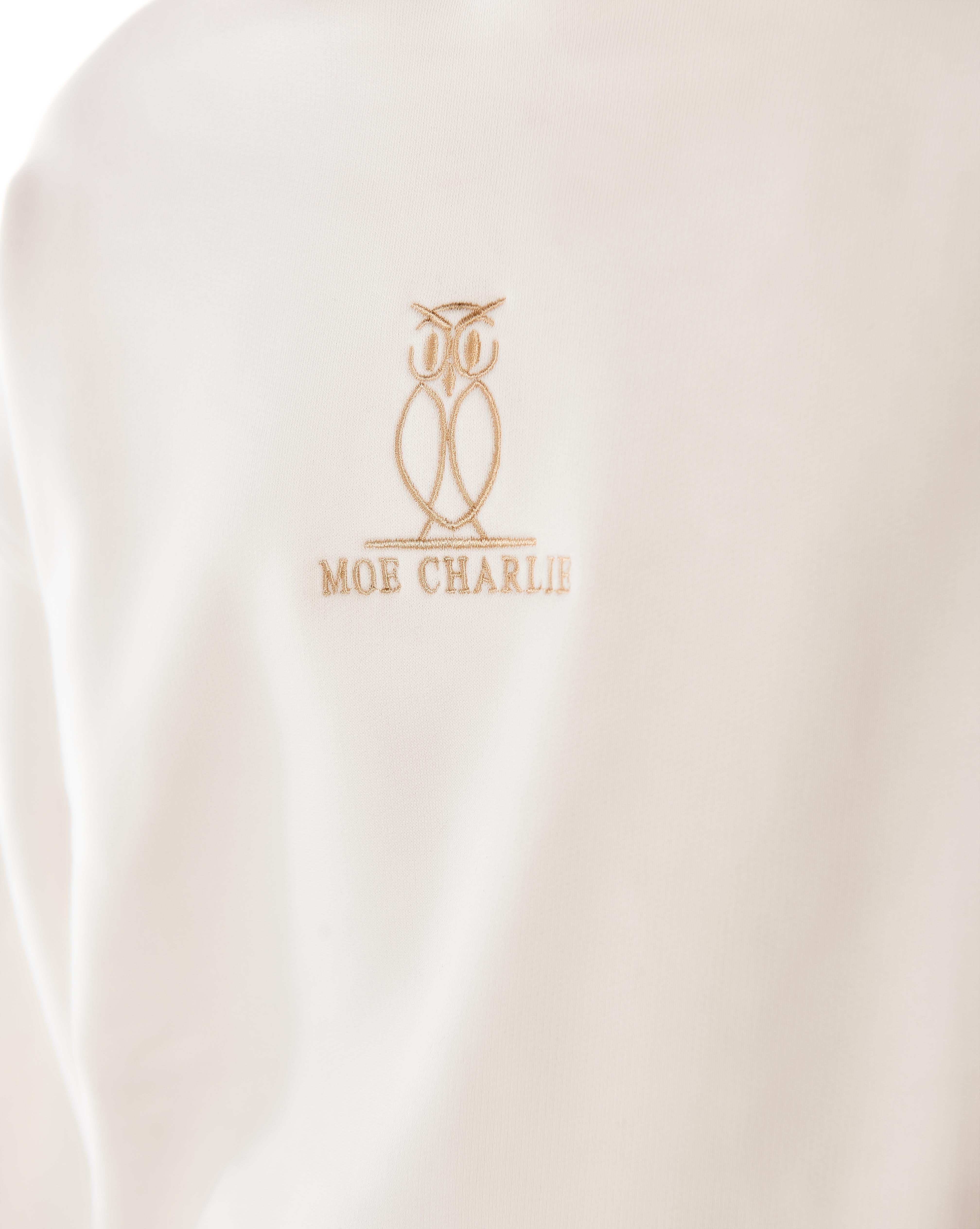 Close-up des goldgestickten MOE CHARLIE Owl-Logos auf dem cremeweißen Star White Hoodie, Detailaufnahme Brustbereich