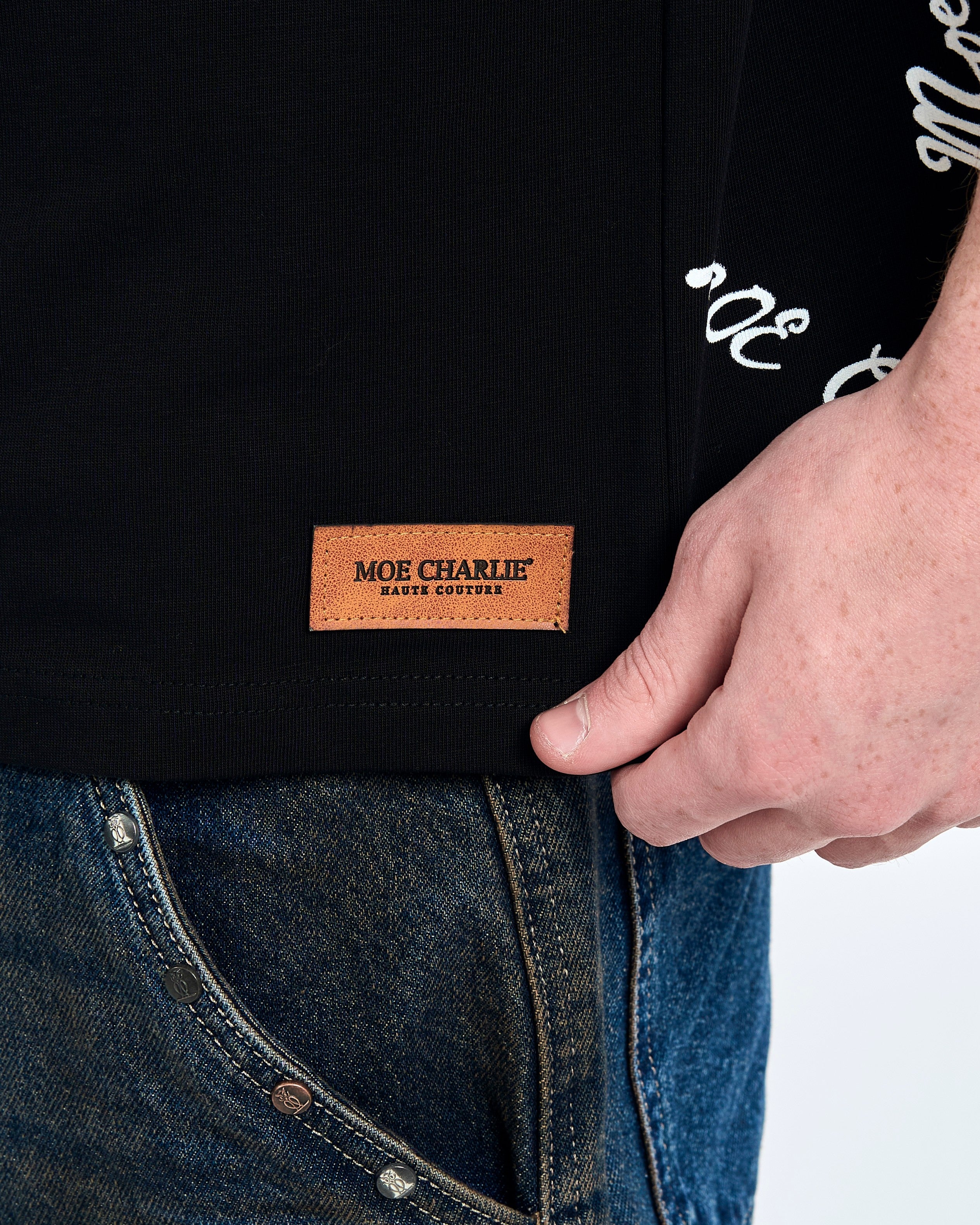 Nahaufnahme Saum - MOE CHARLIE Haute Couture Lederpatch am Forbidden T-Shirt, All-Over-Text im Hintergrund