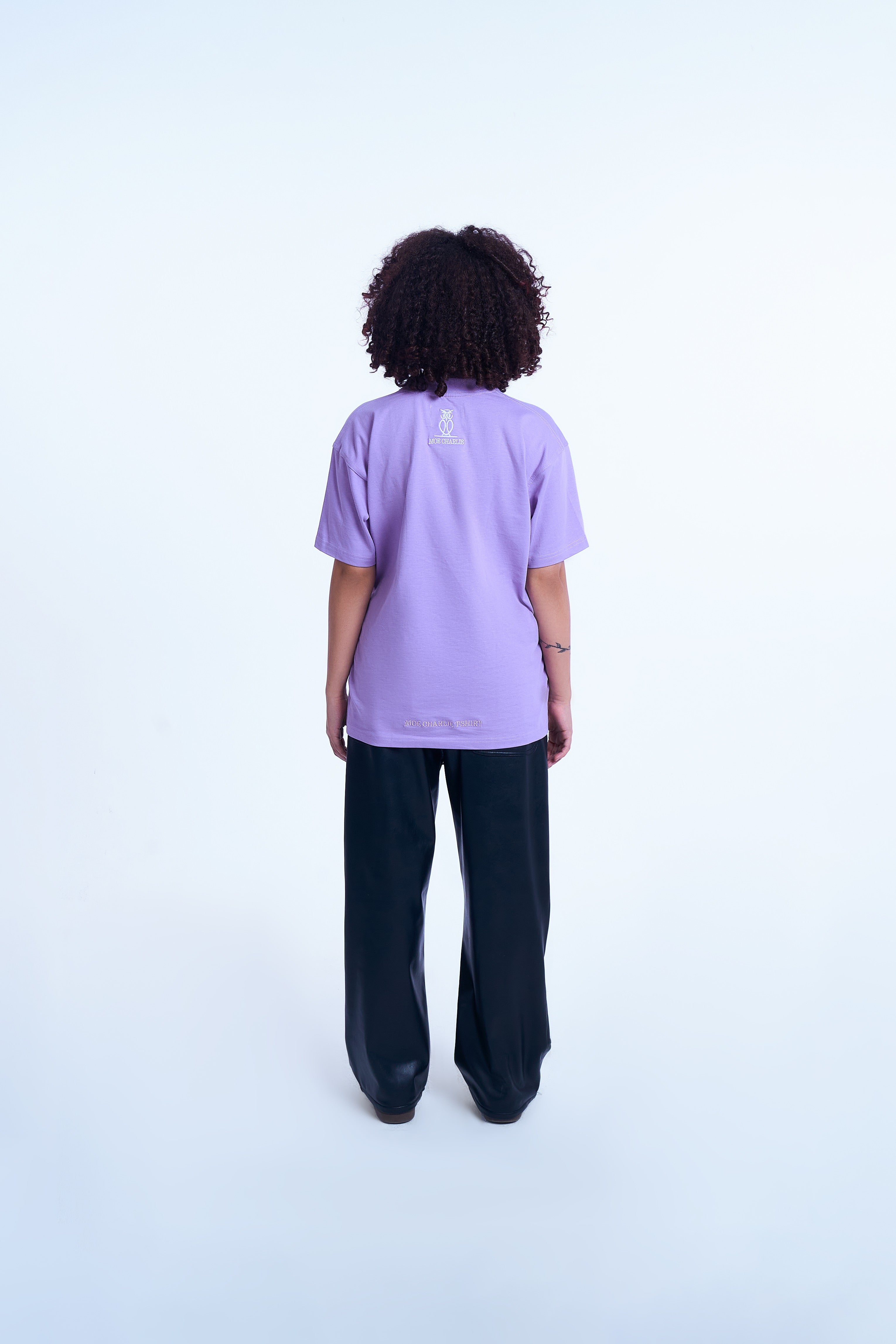 Rückenansicht weibliches Model MOE CHARLIE Basic Tee Lavendel – Eulen-Stickerei am Rücken, schwarze Vinyl-Pants