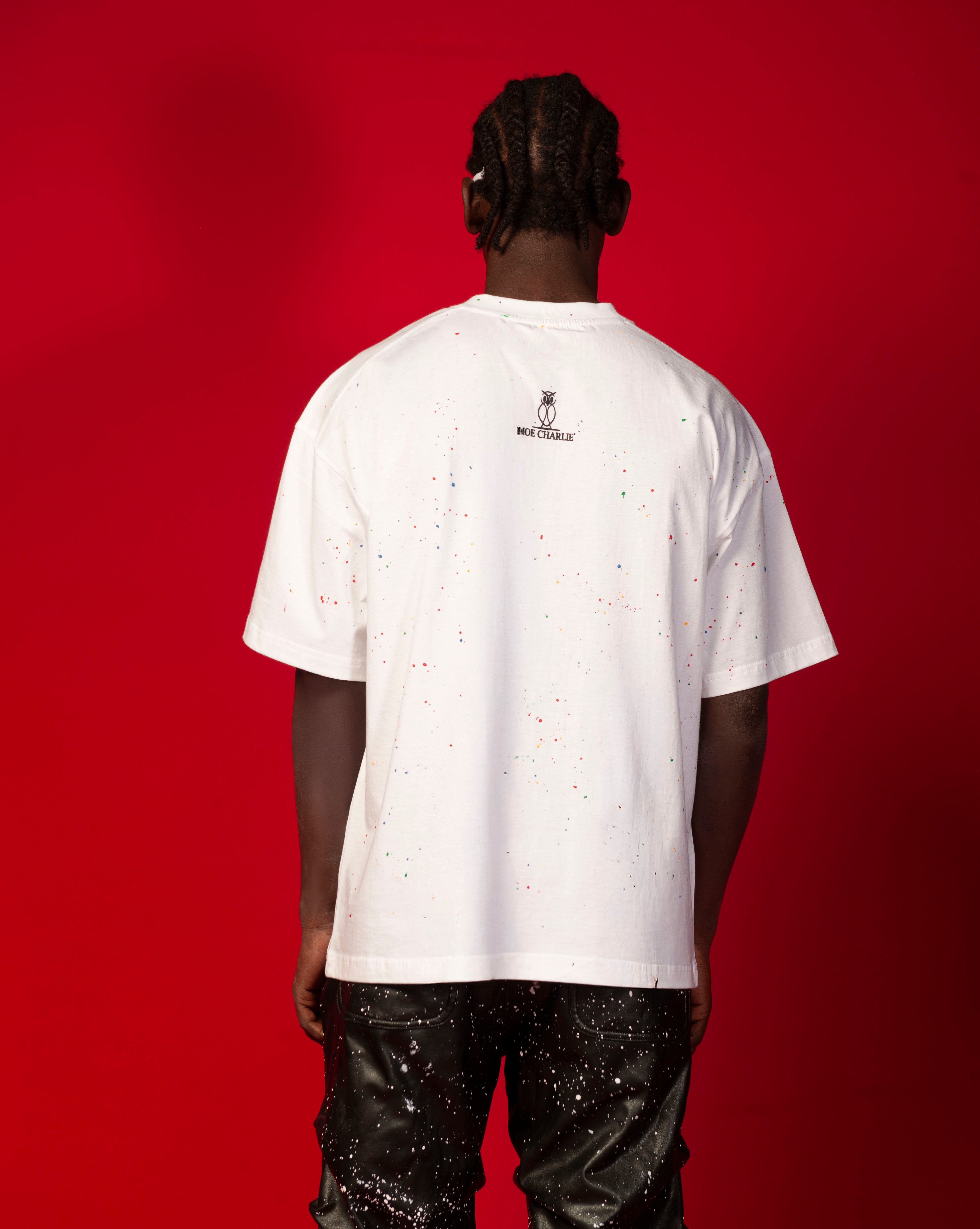 Artist T-Shirt Weiss MOE CHARLIE Rueckenansicht, schwarzes Logo oben, Farbspritzer-Print, dunkle Lederhose kombiniert