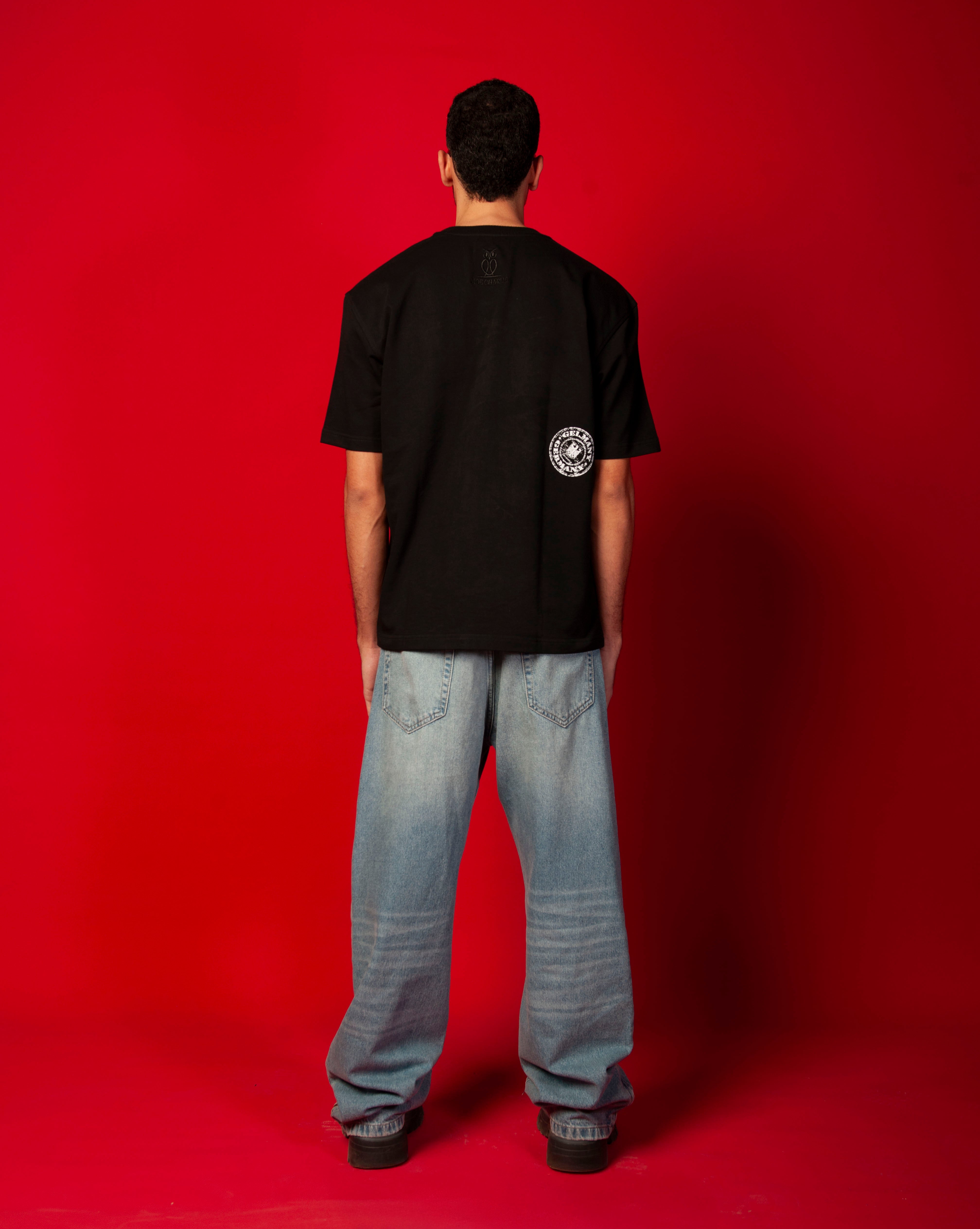 Stampel T-Shirt MOE CHARLIE Rueckenansicht Ganzkoerper, distressed weisser Rund-Stempel auf schwarzem Tee, helle Jeans
