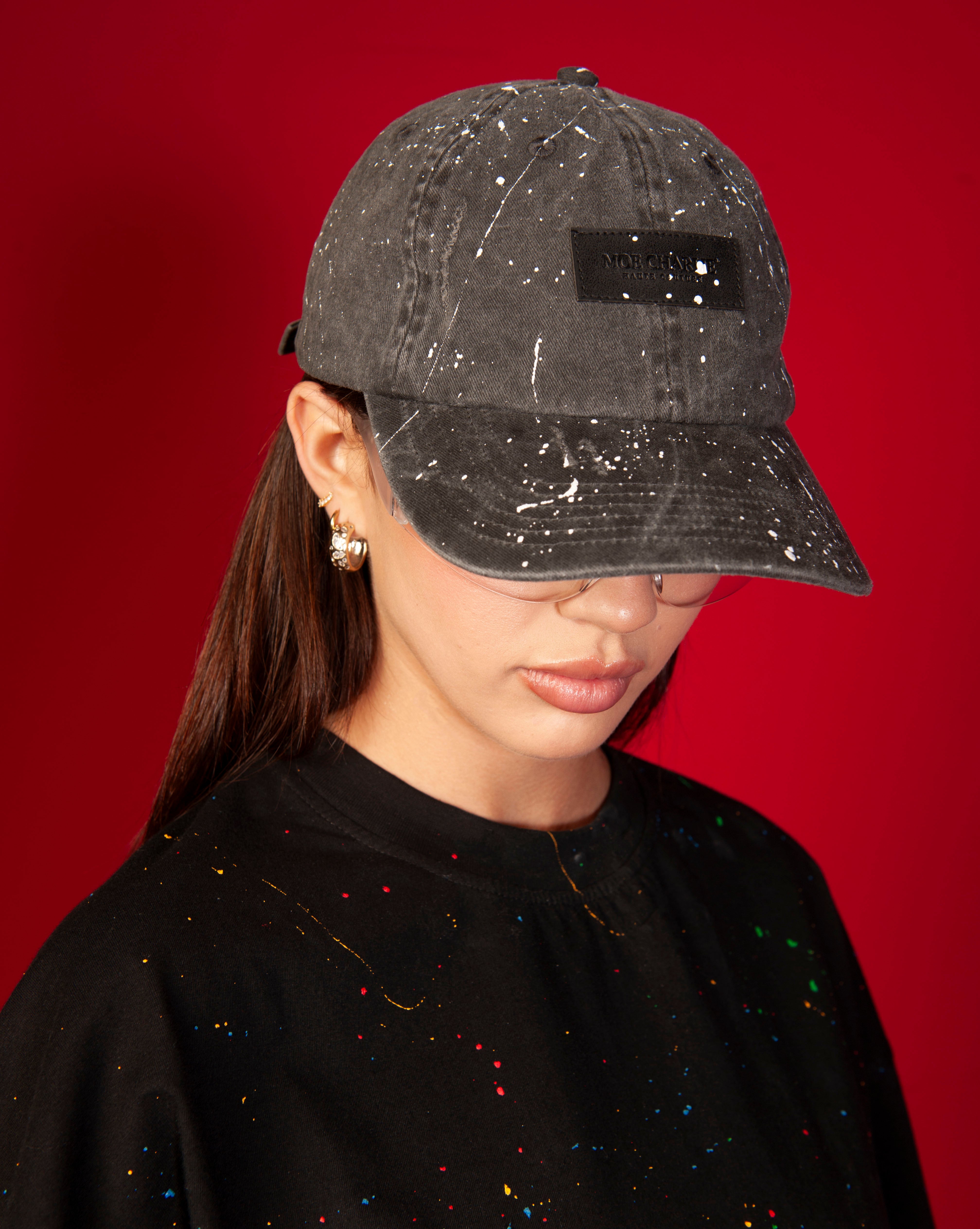 Destroyed MOE CHARLIE Cap, weiblich mit Ponytail, schwarzes Cap tief ins Gesicht gezogen, buntes Farbspritzer-Tee