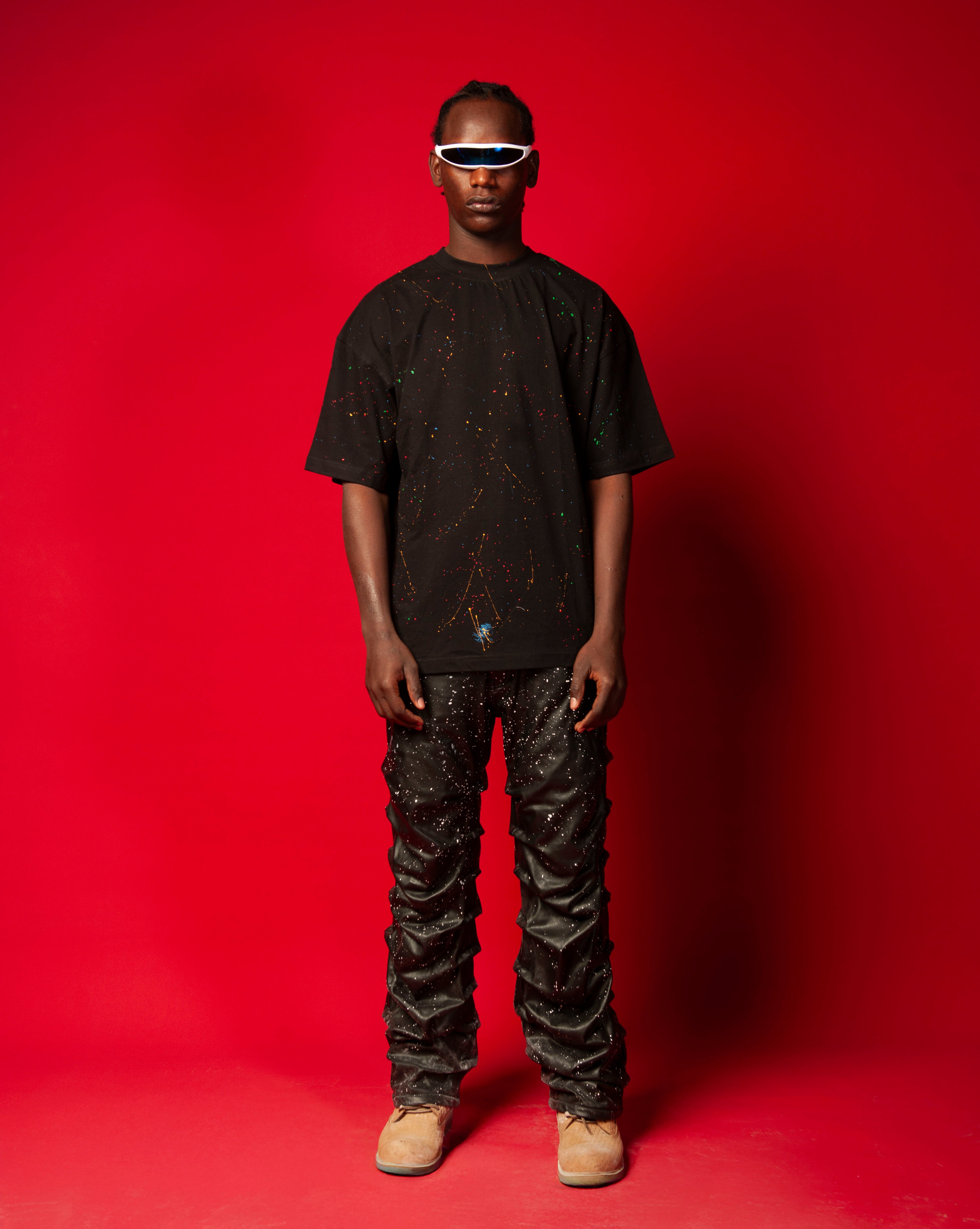 Artist T-Shirt Schwarz MOE CHARLIE, Frontansicht Ganzkoerper, maennlich mit Cornrows, Farbspritzer-Print und Lederhose