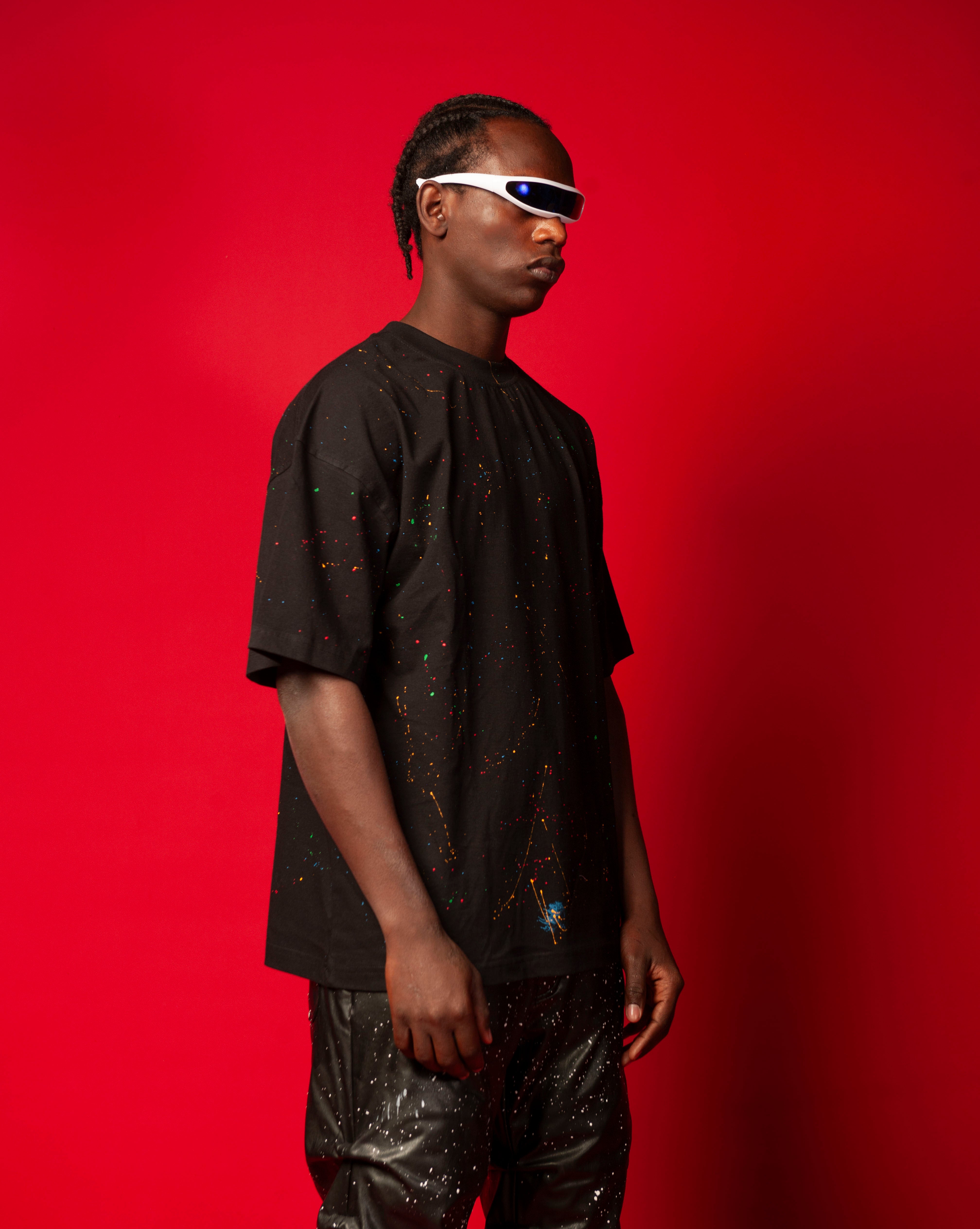 Seitenansicht Artist T-Shirt Schwarz MOE CHARLIE, maennlich mit Cornrows, bunter Farbspritzer auf schwarzem Oversize-Tee