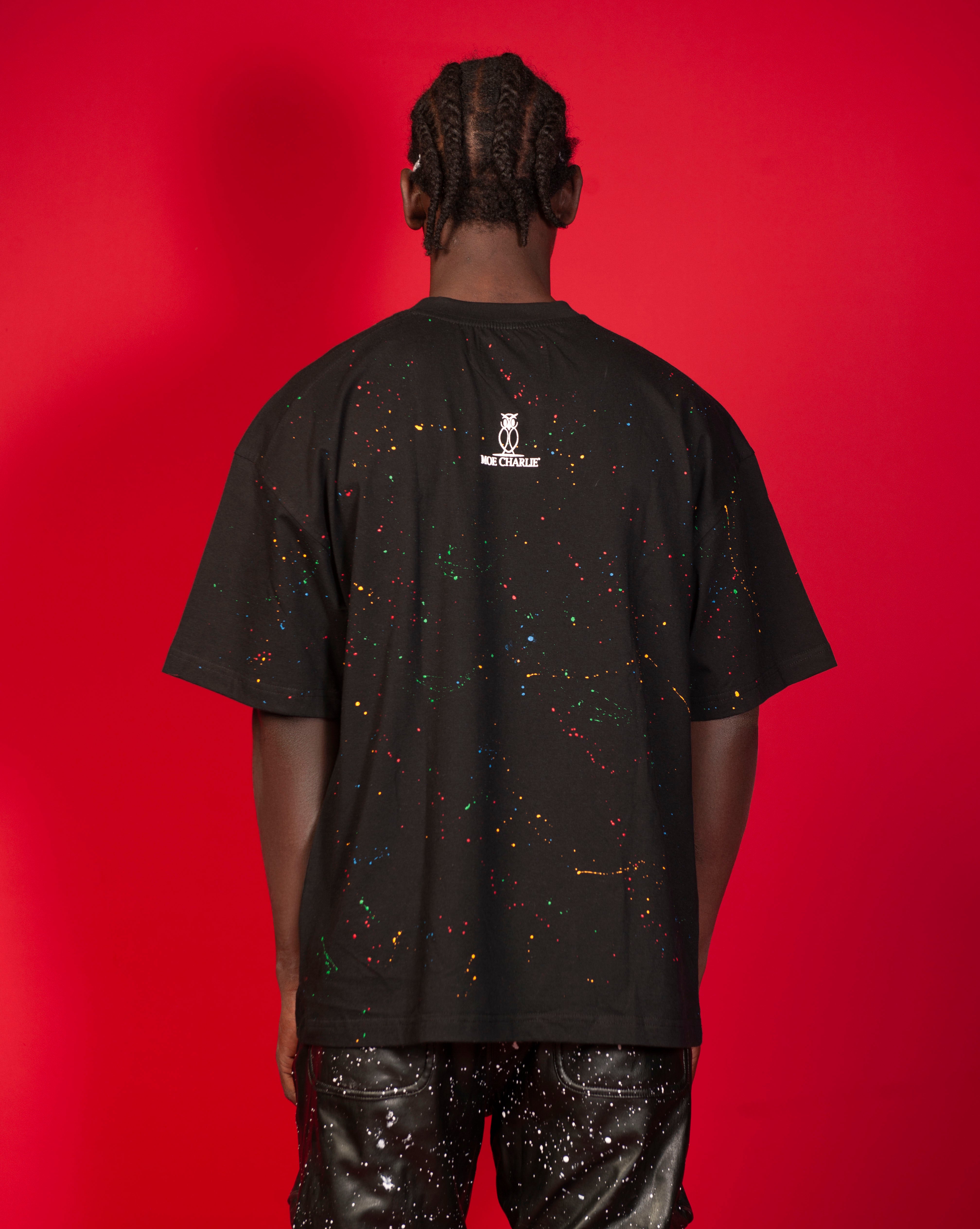 MOE CHARLIE Artist T-Shirt Schwarz Rueckenansicht Ganzkoerper, maennlich mit Cornrows, schwarze Lederhose mit Spritzer