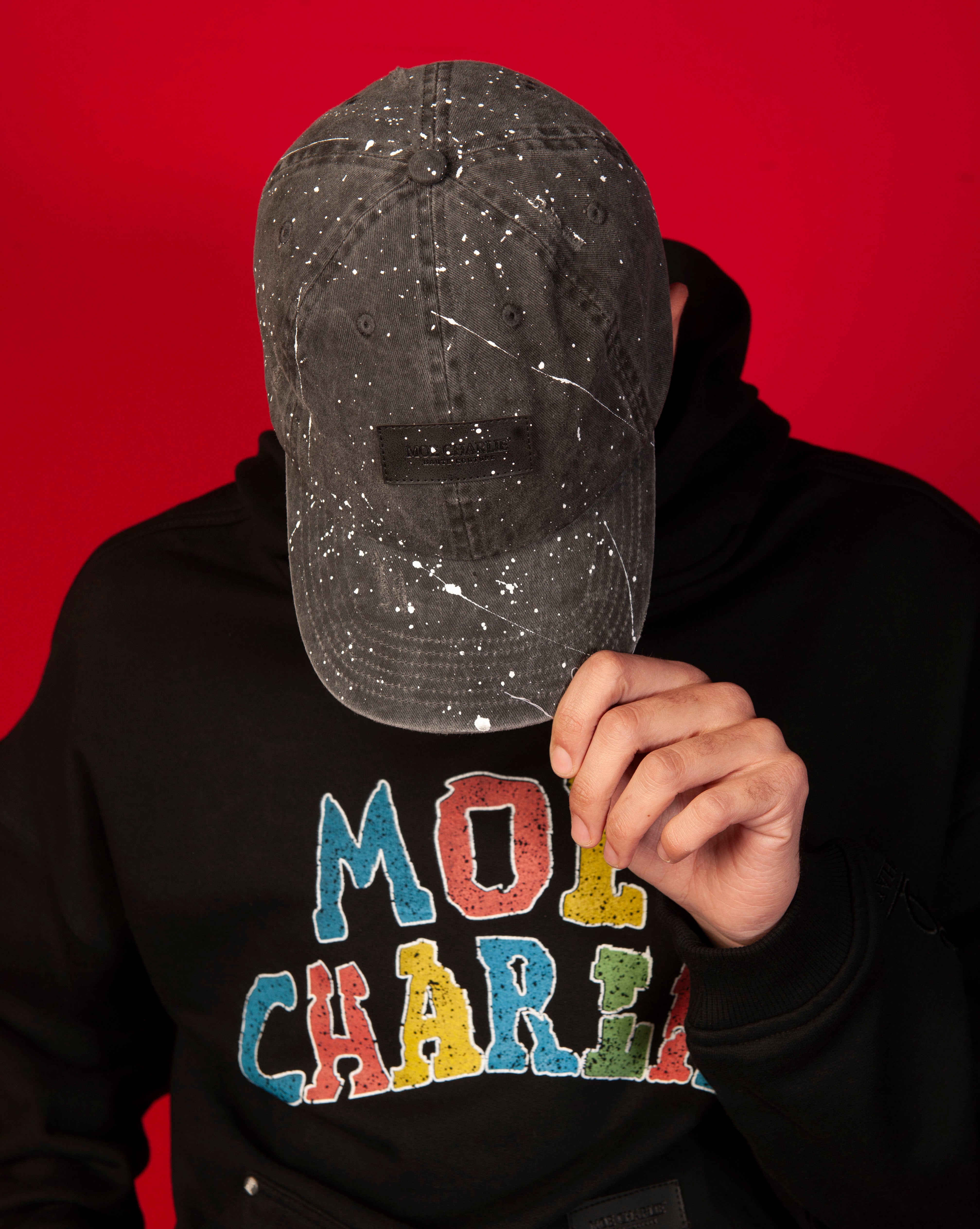 MOE CHARLIE Destroyed Cap von vorne, maennlich, Cap haelt Gesicht verdeckt, schwarzes Farbspritzer-Cap mit Label