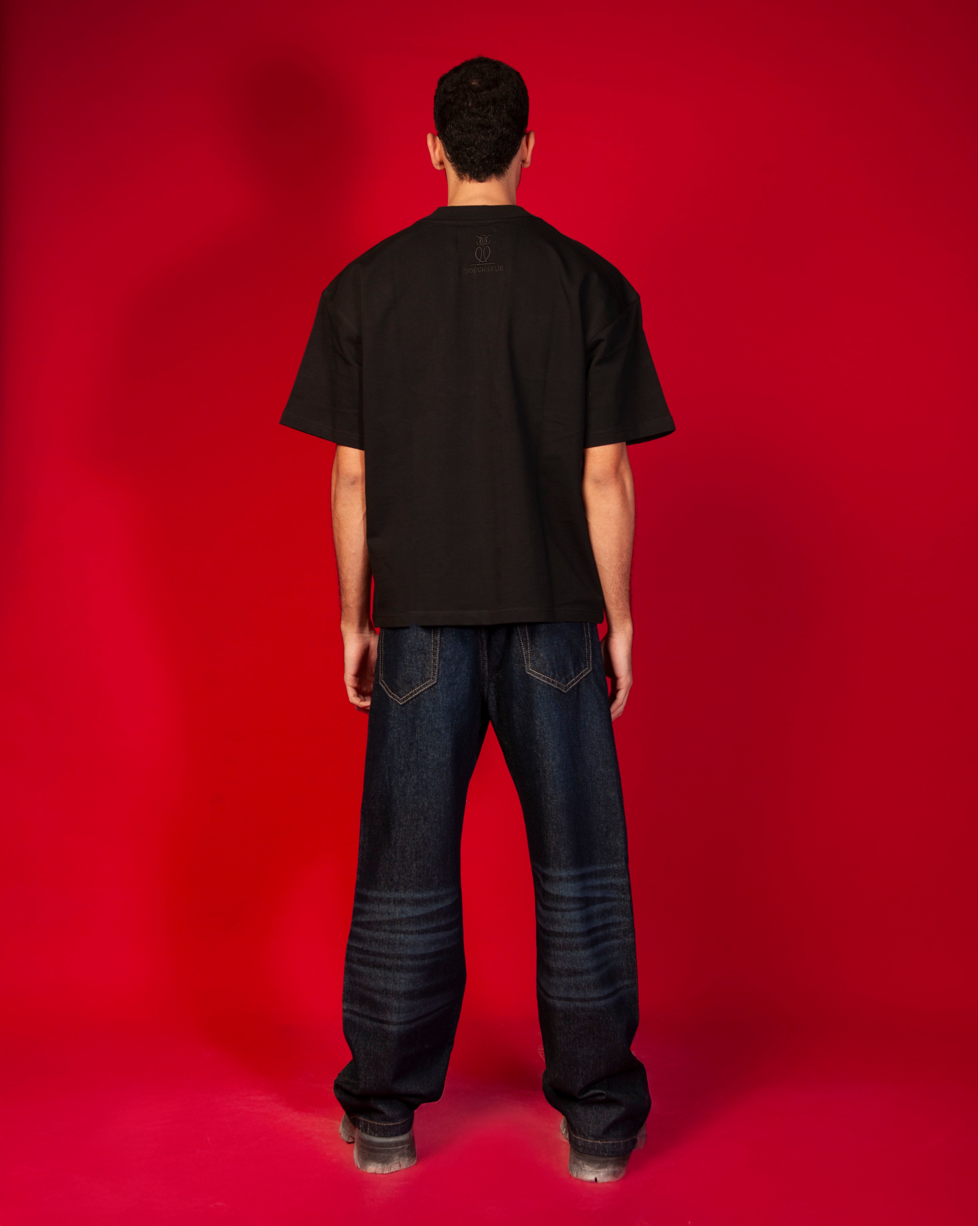 Rueckenansicht MOE Oversize T-Shirt MOE CHARLIE, schwarze Rueckseite mit kleinem MOE CHARLIE Logo oben, dunkle Jeans